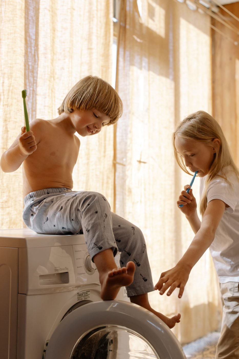 Dos niños lavándose los dientes sentados sobre la lavadora, junto a una ventana con luz de mañana.