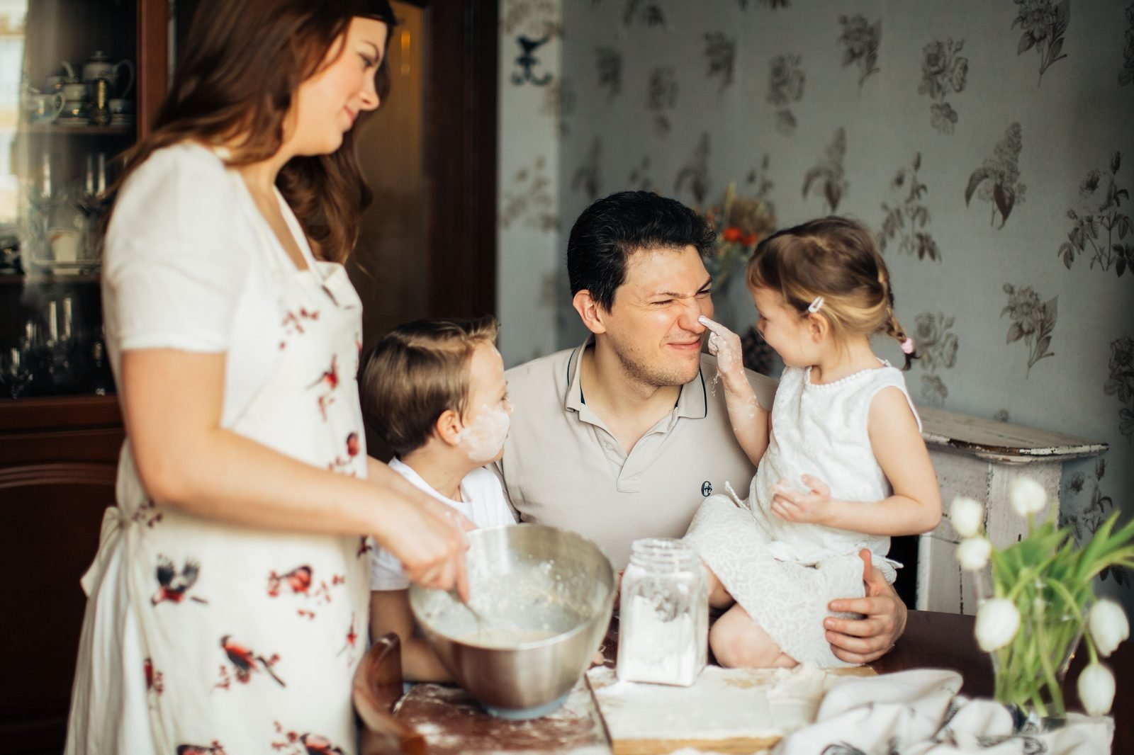 Familia preparando la comida en la cocina: una adulta con un bol mientras un adulto sostiene a una niña y un niño observa.